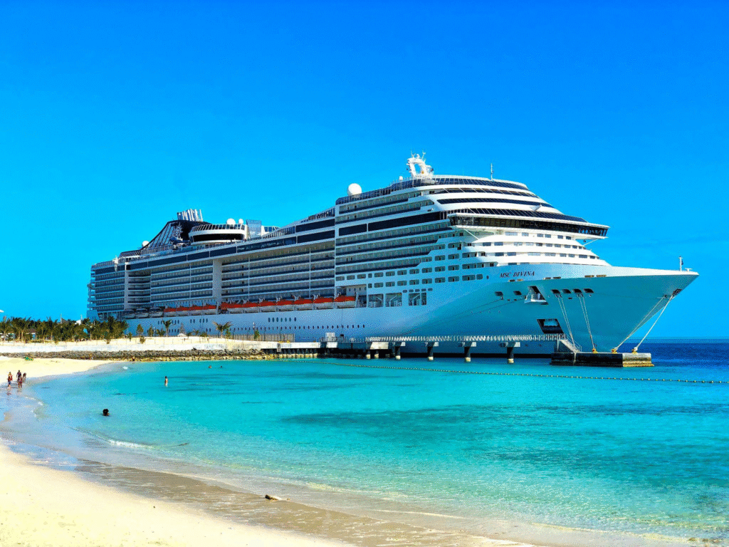 A cruise ship docked by the shore with a clear blue sky and beach in the foreground.